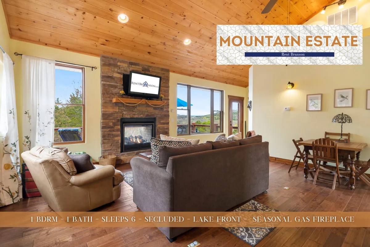 A warm, open-plan living area featuring a seasonal gas fireplace, plush seating, large windows framing stunning views of the lake and woods, and a stylish dining area adjoining the living space. The room exudes comfort with cozy furnishings and natural light flooding in, making it perfect for gatherings or quiet evenings.