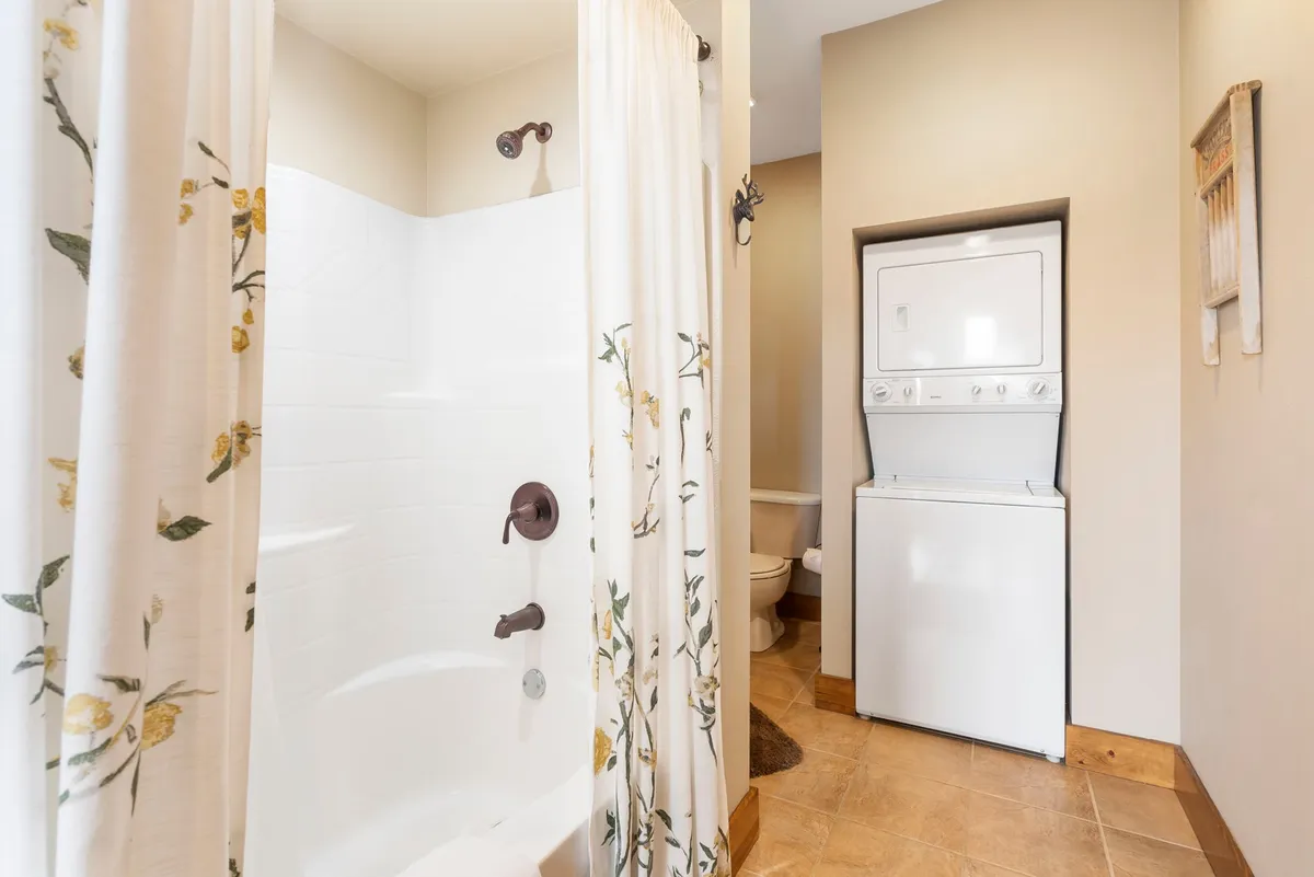 A functional bathroom highlighted by a shower area with a floral curtain, stacked washer and dryer, and soothing neutral-colored walls. The space is compact yet welcoming, with convenience and relaxation in mind.