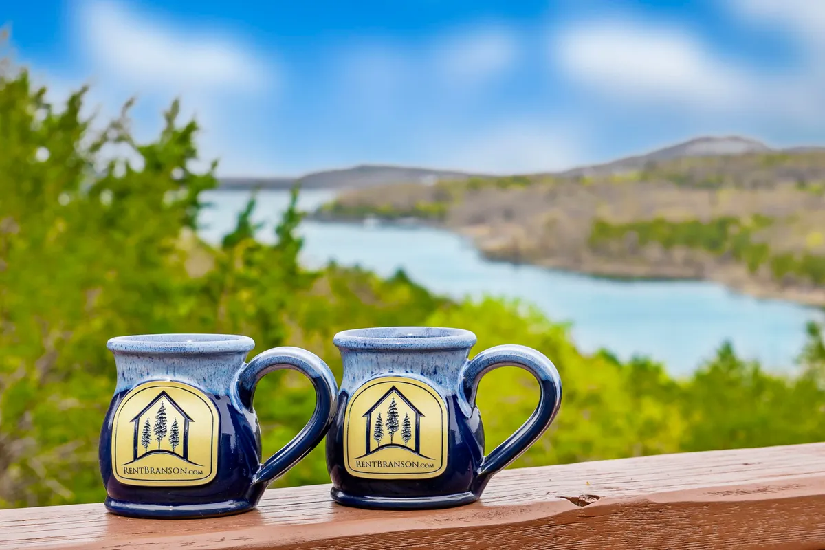 This image features two mugs resting on the railing of a deck, with a lush green landscape and the sparkling blue lake visible in the distance. Sunlight filters through the trees, creating a serene setting that invites moments of quiet relaxation and connection with nature.