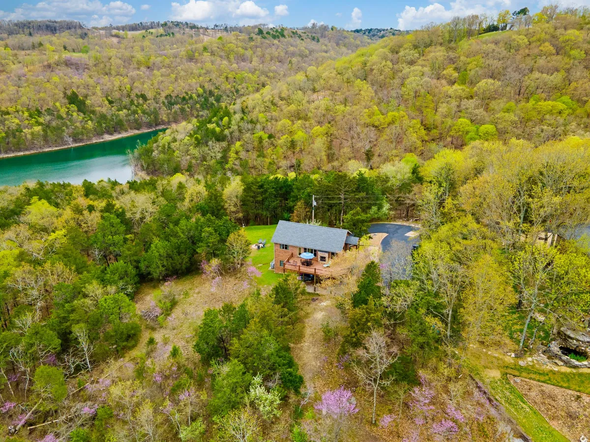 The image showcases a charming single-family home nestled among towering trees, overlooking the shimmering waters of Table Rock Lake. A winding path leads from the house through wildflowers toward the water’s edge, with a peaceful sunset casting a golden glow over the entire scene. The expansive natural setting evokes seclusion, tranquility, and opportunities for outdoor adventure.