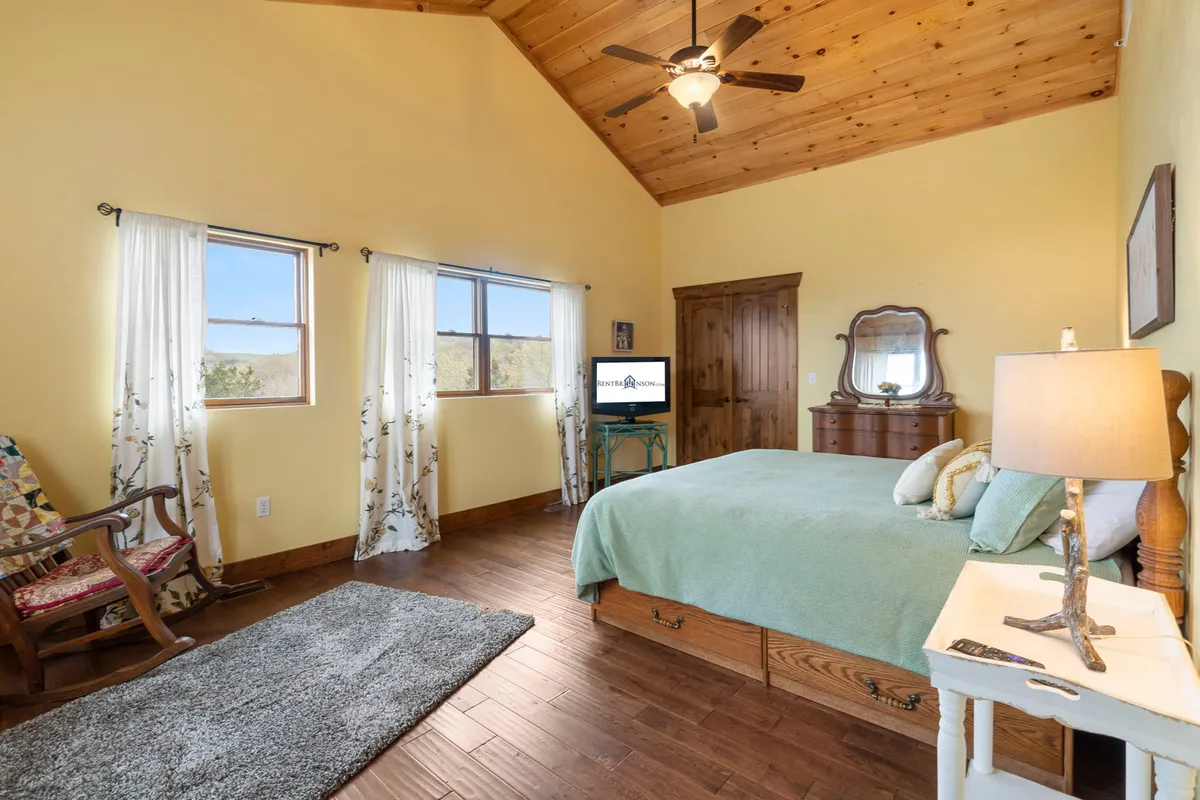 A tranquil bedroom with a bed dressed in a light green blanket, exposed wooden ceiling beams, windows letting in gentle natural light, and a cozy rocking chair in the corner. The room evokes a peaceful, restful atmosphere, perfect for unwinding.