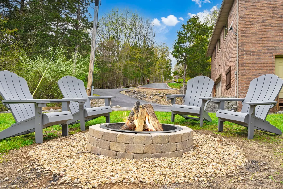 A cozy fire pit surrounded by several Adirondack chairs, set against a backdrop of tall trees and natural greenery. The setup suggests a welcoming, communal space for gathering, storytelling, and enjoying evenings under the stars.