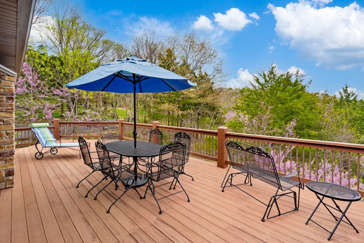 A serene wooden deck with a large blue umbrella and a black metal dining set surrounded by greenery. The inviting setup suggests relaxation, outdoor meals, and opportunities for solitude or gentle social interaction with nature as a backdrop.