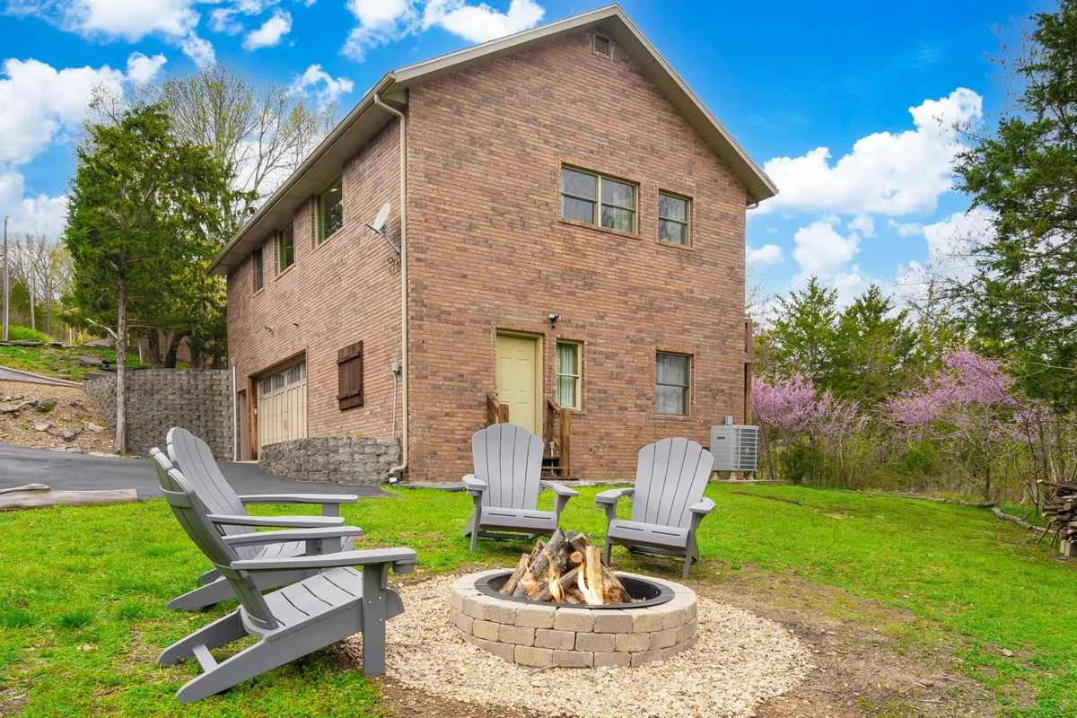 This exterior scene shows a cozy fire pit area with Adirondack chairs set around it, brick house in the background, and surrounding greenery. The ambiance suggests intimate fireside moments and the opportunity to enjoy the outdoors in comfort and warmth.