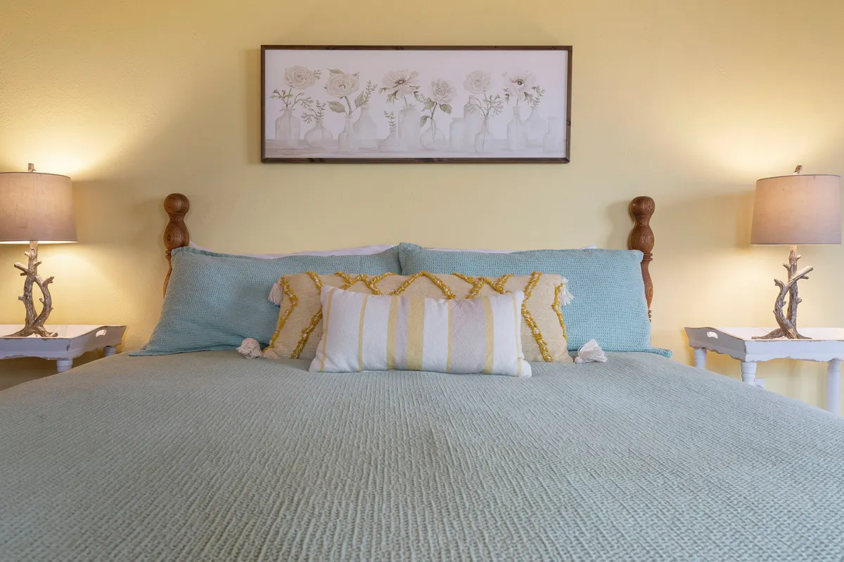 A bright, comforting bedroom with a plush bed featuring textured bedding, floral artwork above the headboard, and matching nightstands with lamps. Sunlight streams in, enhancing the inviting and restful mood.