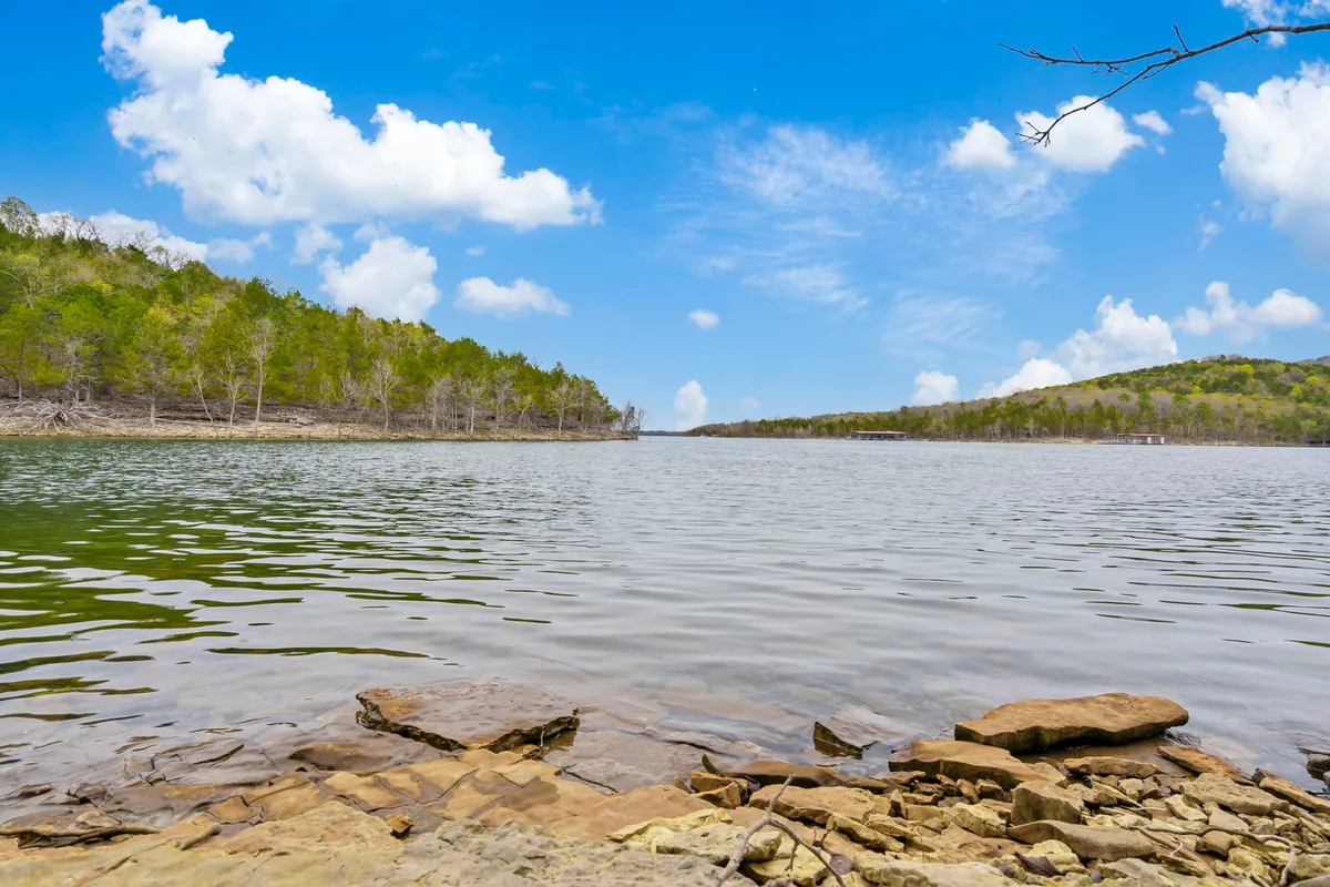 A wide, tranquil view of Table Rock Lake stretches out, with calm water, green hills, and a rocky foreground. The scene is peaceful, inviting viewers to imagine quiet moments by the water’s edge, watching sunsets or taking starlit strolls.