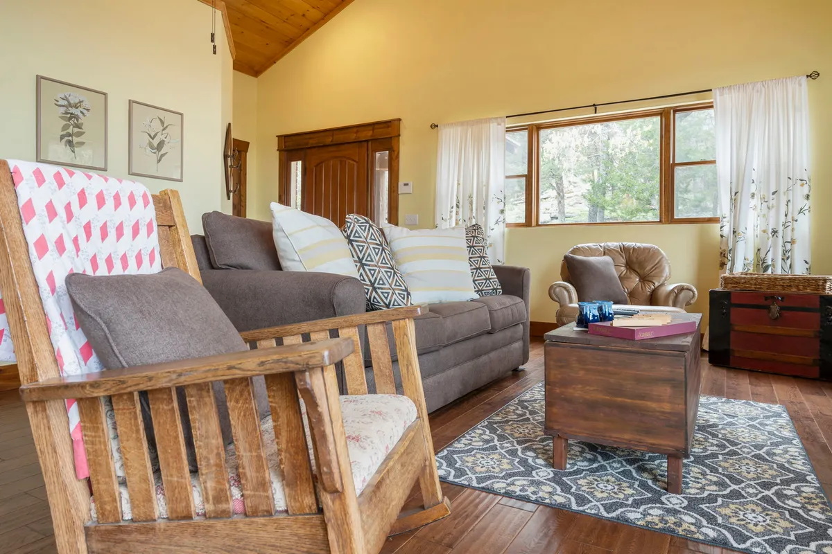 A comfortable seating nook in the living space, featuring a sun-filled window, a plush sofa, a wooden coffee table with books, and a classic rocking chair. The ambiance is bright and restful—ideal for curling up with a book, enjoying morning coffee, or sharing stories with loved ones.
